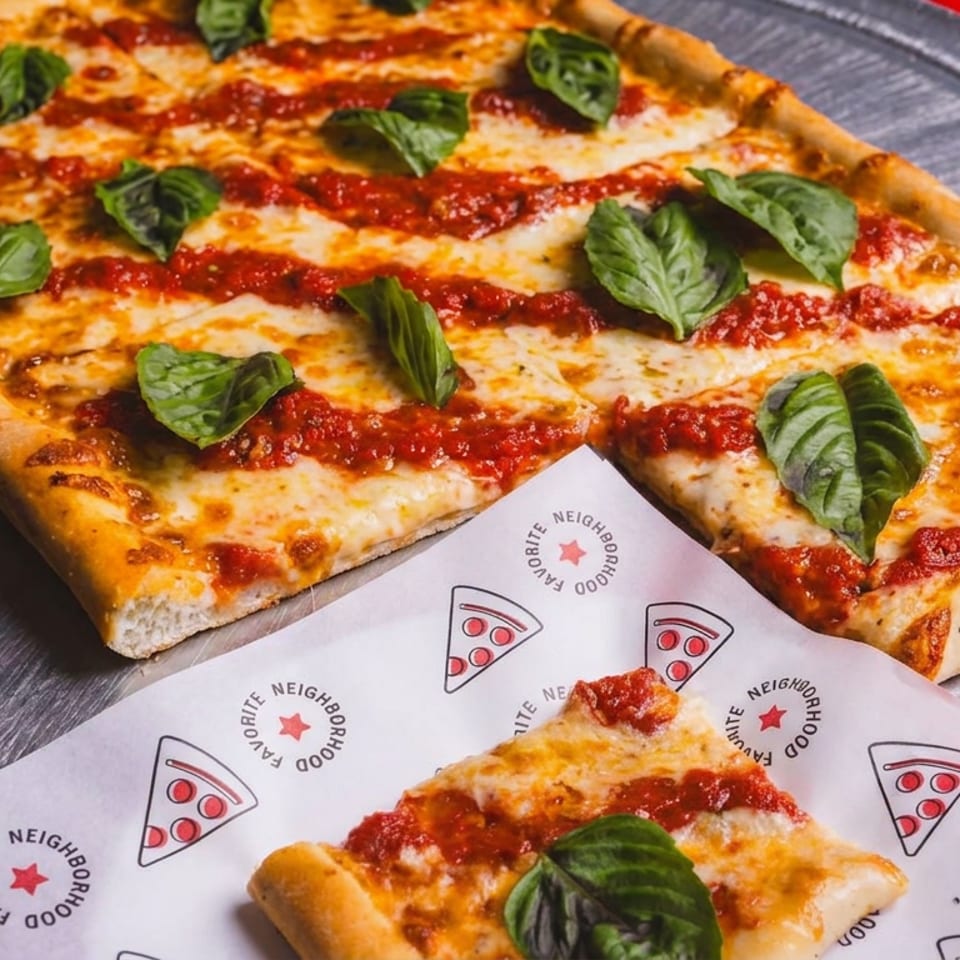 Rectangular cheese pizza topped with tomato sauce and fresh basil sits on a metal tray beside a branded slice.