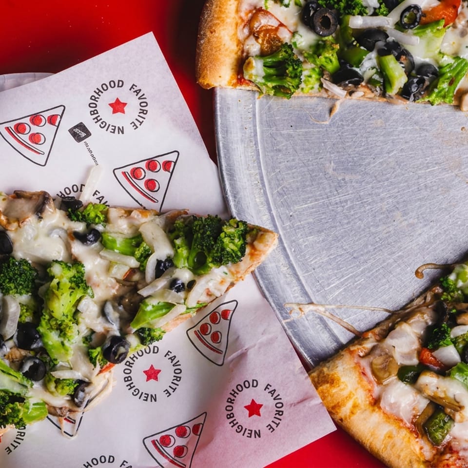 Three slices of vegetable pizza with broccoli, mushrooms, onions, peppers, black olives, and melted cheese on branded paper and a metal tray.