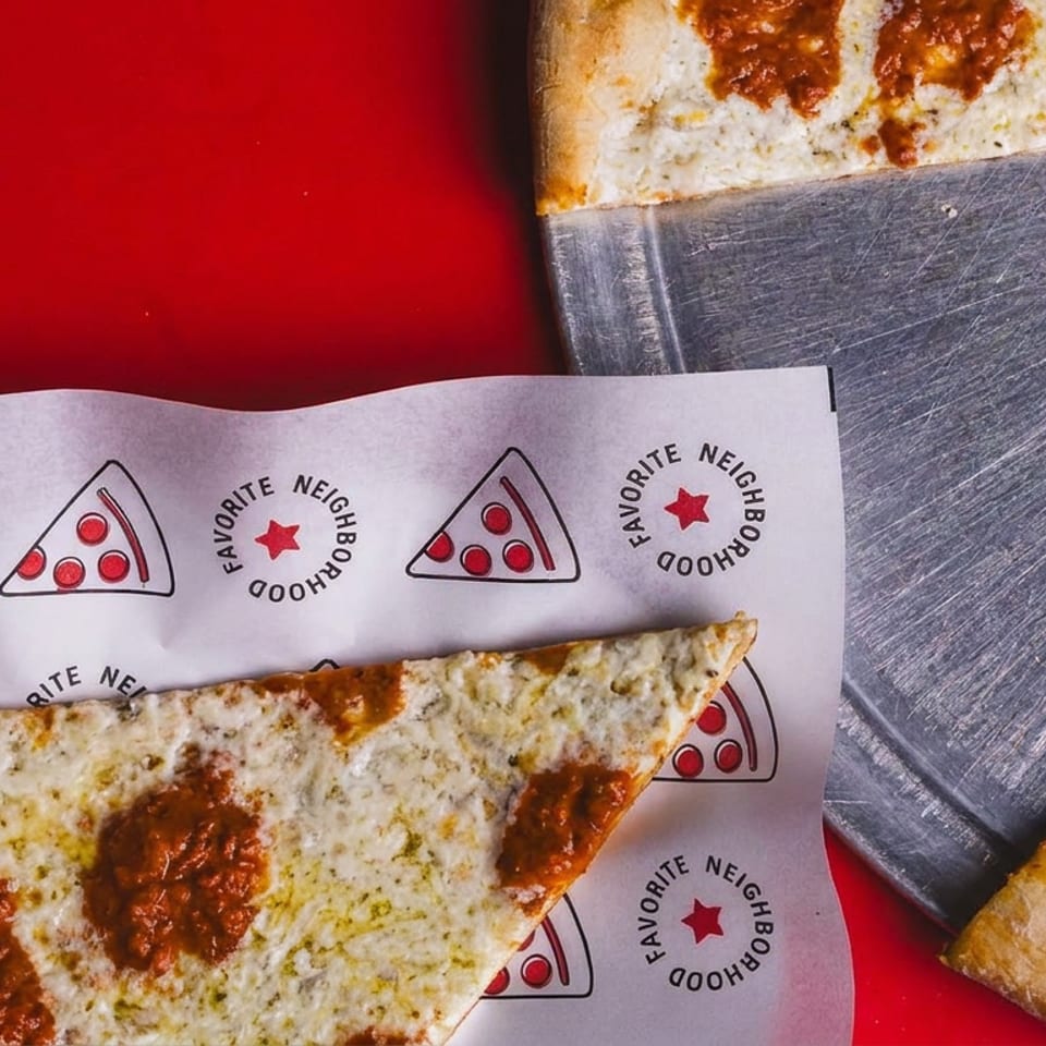Two cheesy pizza slices with pepperoni sit on branded paper beside a metal serving tray against a red background.